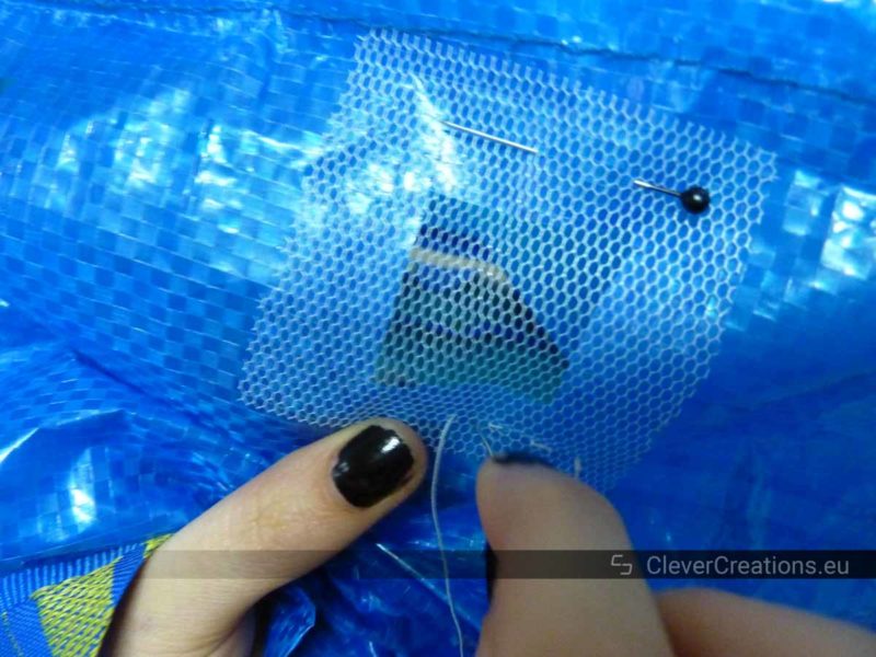 A close-up of a pair of hands using a sewing needle to sew a square piece of window mesh to an IKEA bag.
