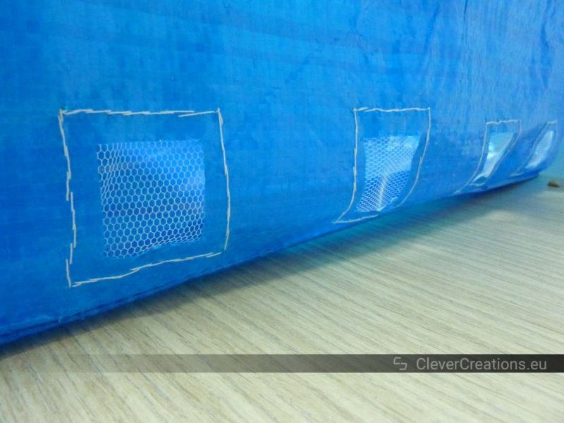 The outside of an IKEA FRAKTA bag with four drainage holes that are covered by window mesh.