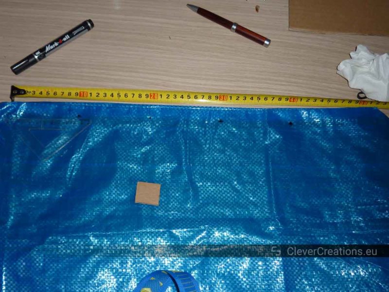 An IKEA FRAKTA bag with dotted markings, next to it an extended measuring tape and some pens.