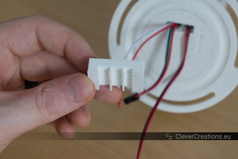 A hand holding a 3D printed white strain relief component in front of a white plastic part with wires extending from it.