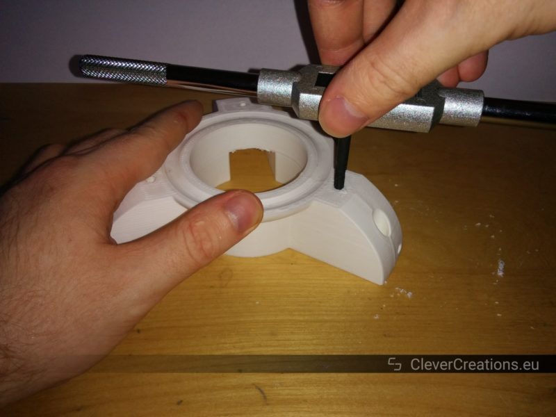 A hand holding a M5 threaded tap that is being used to tap thread in a hole in a white 3D printed component that is held by another hand.