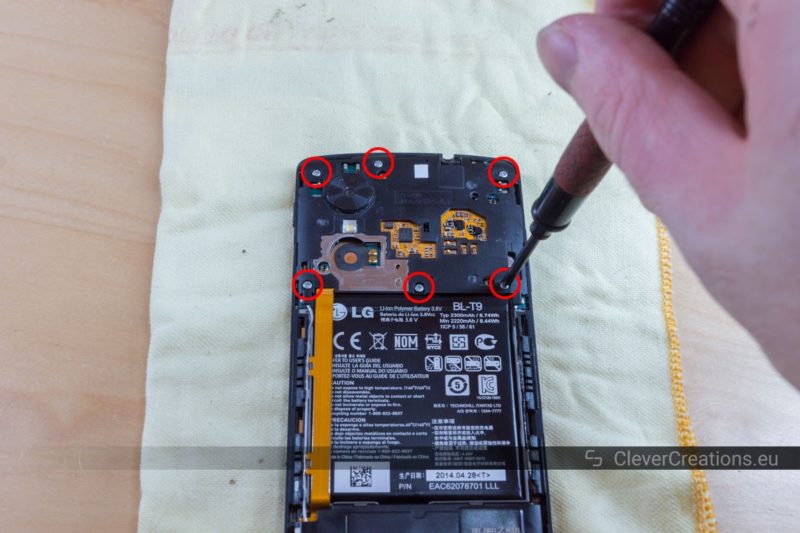 A hand holding a screwdriver and removing one out of six circled screws on the motherboard cover of a Nexus 5 phone.