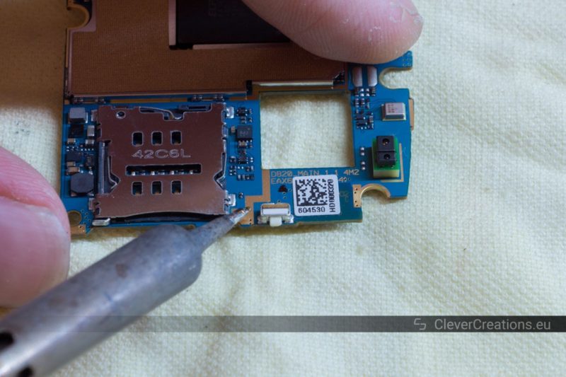 A soldering iron pointing at the power button of a Nexus 5 motherboard.
