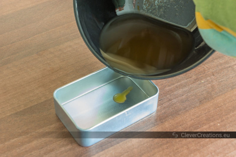 A liquid wax mixture poured from a black bowl into a tin storage container.