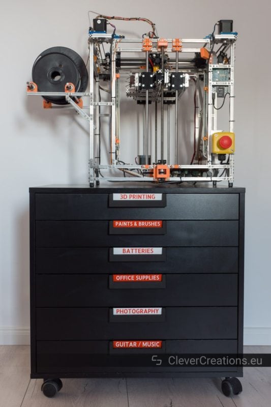 A single black IKEA ALEX drawer unit on wheels, with 3D printed handles and red labels, and on top of the drawer unit a 3D printer.