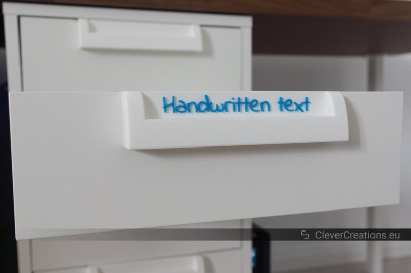 An extended drawer on a white IKEA ALEX drawer unit, with a 3D printed handle and white label with blue text.