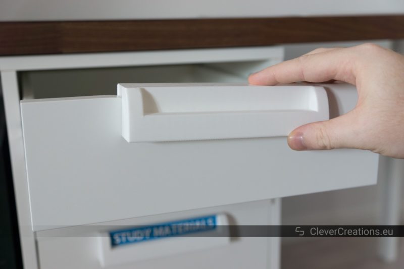 A hand placing a white 3D printed handle on the extended drawer of an IKEA ALEX unit.