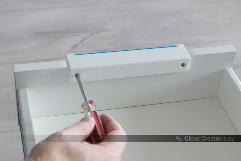 A hand using a screwdriver to drive a screw into an IKEA ALEX drawer to secure a 3D printed handle.