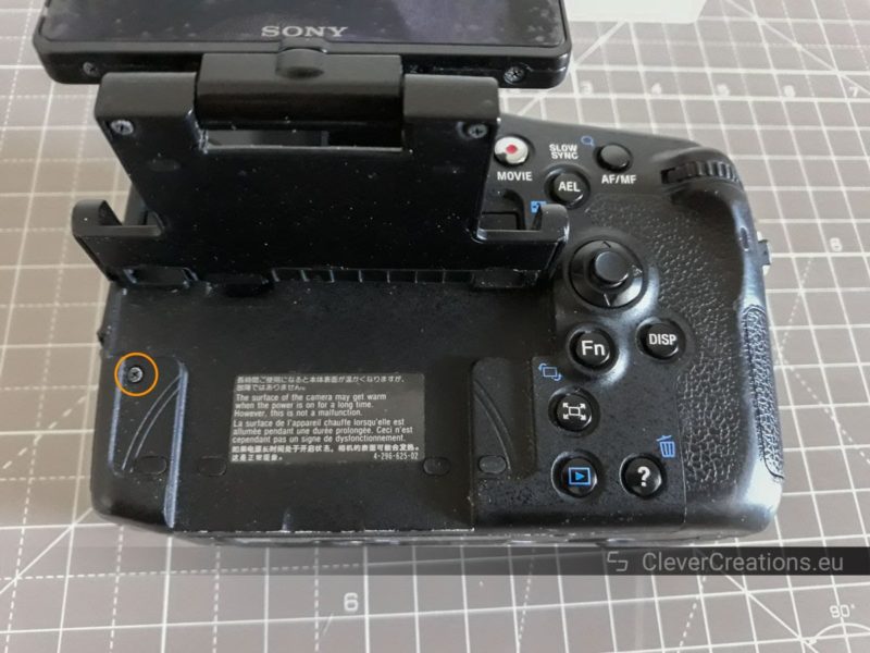 The back of a Sony SLT-A77 camera body with a lifted LCD screen. One exposed screw is circled.