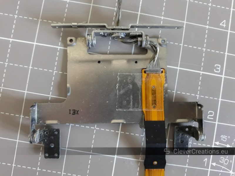 A metal hinge assembly and a flex cable that has been attached to it using clear tape.