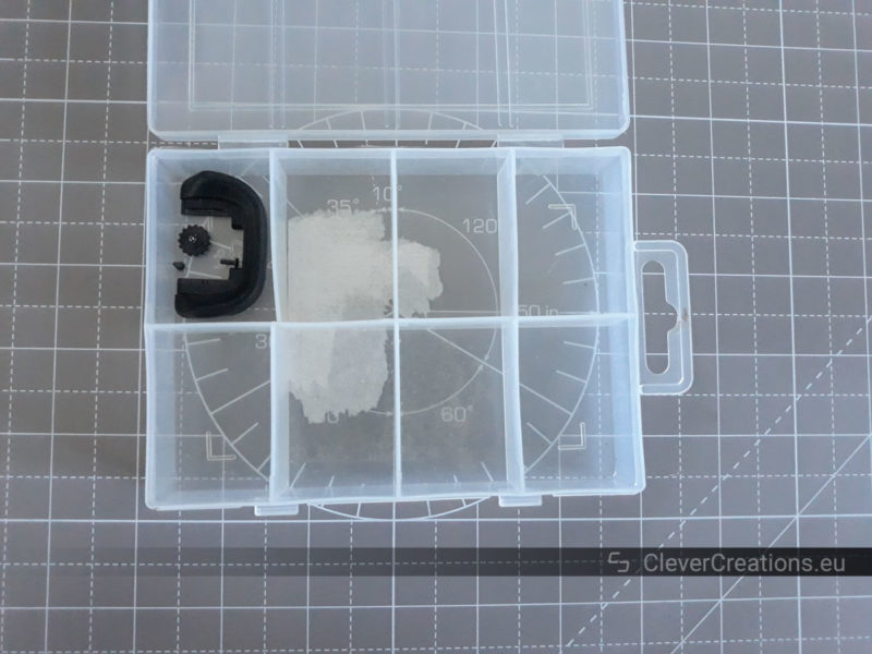 Top view of a small transparent box with separators that contains some screws and a rubber eye cup from a camera.