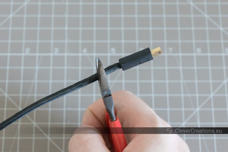 A hand holding a pair of pliers that is used to cut through a braided USB cable with USB mini-B connector.