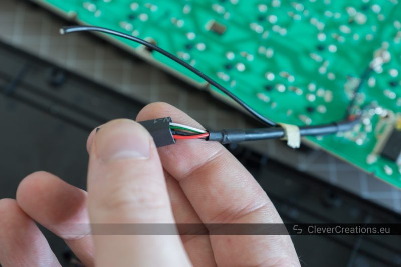 A hand sliding a piece of heat shrink tubing over exposed wires of a USB cable. In the background a large PCB of a computer keyboard.