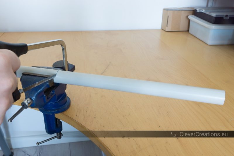 A hand using a hacksaw to cut a grey PVC pipe that is clamped in a vise.