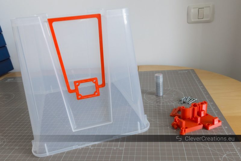 An upside down 22L IKEA SAMLA box with a red drill jig placed on top, with red 3D printed components next to it.
