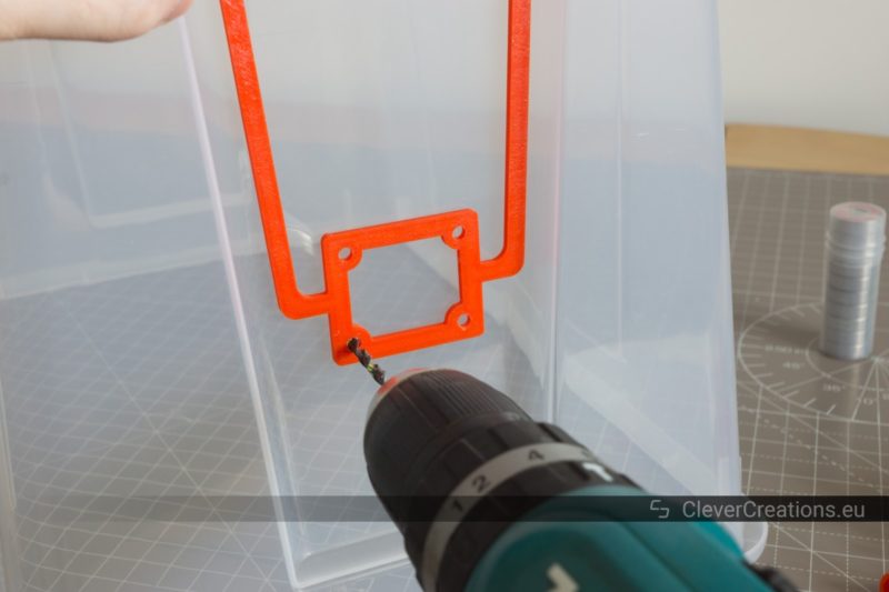 A power drill drilling a hole in a 22L IKEA SAMLA box using a 3D printed red drill jig as a guide.