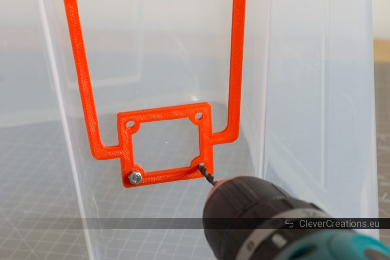 A power drill drilling a hole in a 22L IKEA SAMLA box using a 3D printed red drill jig as a guide.