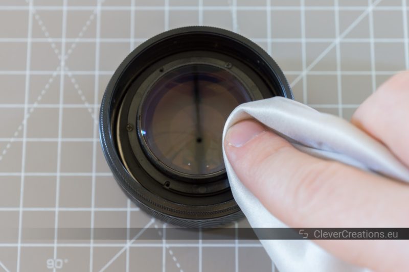 A hand using a microfiber cloth to wipe dust from a lens front element.