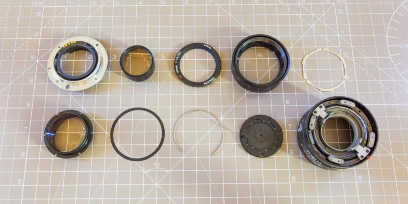 A fully disassembled Minolta Sony SAL50F14 camera lens with all components laid out next to each other on a cutting mat.