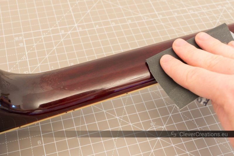 A hand holding a Micro-Mesh sanding sheet and sanding lacquer on the back of an electric guitar neck.