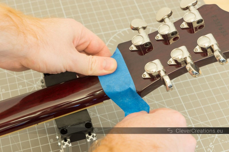Two hands applying blue painter's tape on the headstock of a guitar.