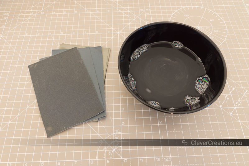 A stack of Micro-Mesh sanding sheets next to a black bowl of soapy water.