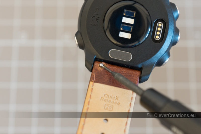 A spring bar tool pointing at the quick-release pin button of a leather watch strap of a Garmin Forerunner 245 watch.
