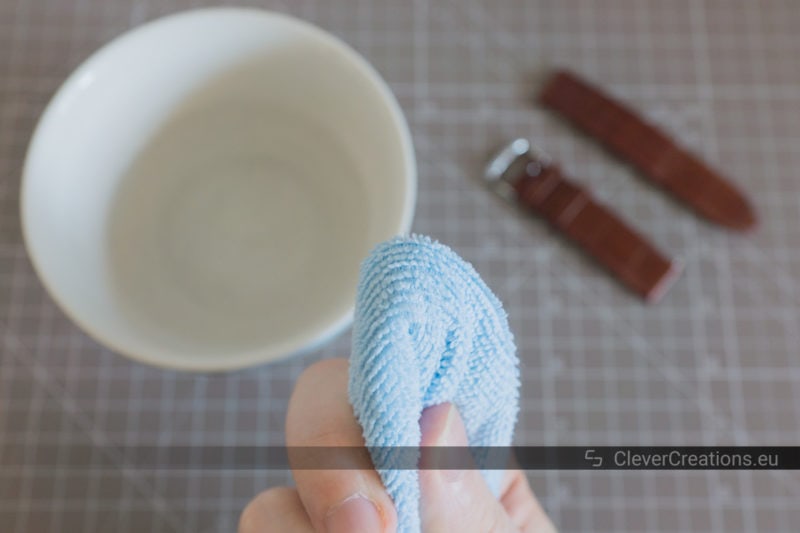 A damp blue microfiber cloth being held in front of a brown leather watch strap and a bowl of water.