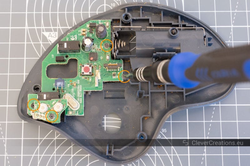 A screwdriver unscrewing a screw on a trackball circuit board, with 4 circled screws.