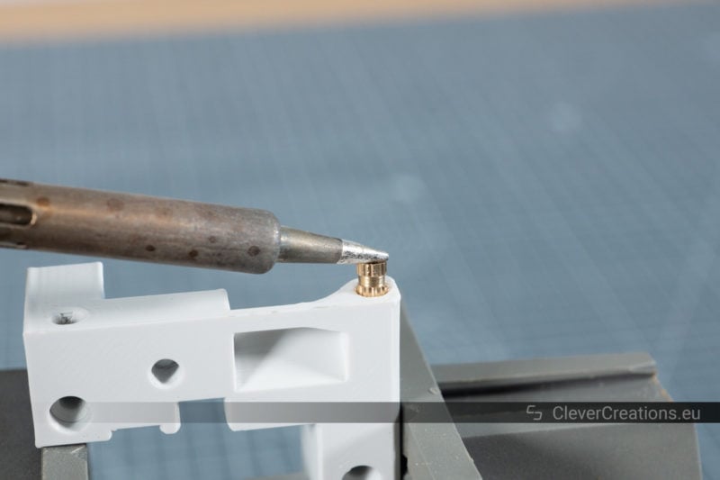 The tip of a soldering iron pressing a brass threaded insert into a hole in a grey plastic part.