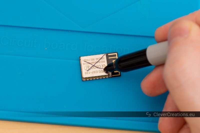 A black marker being used to mark a cross on the top metal cover of an ESP8266 module.
