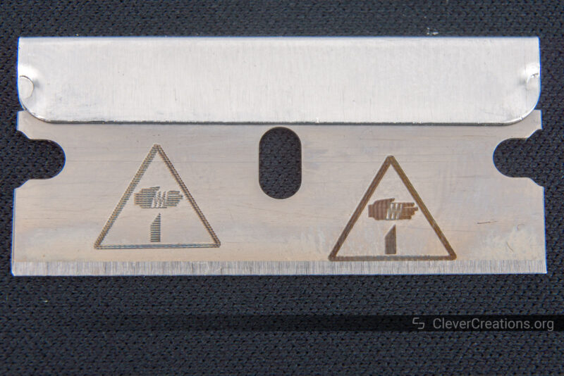 A stainless steel razor blade with two 'danger' symbols engraved on its surface