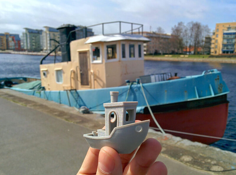 A 3D printed benchy held in front of a boat in a river.