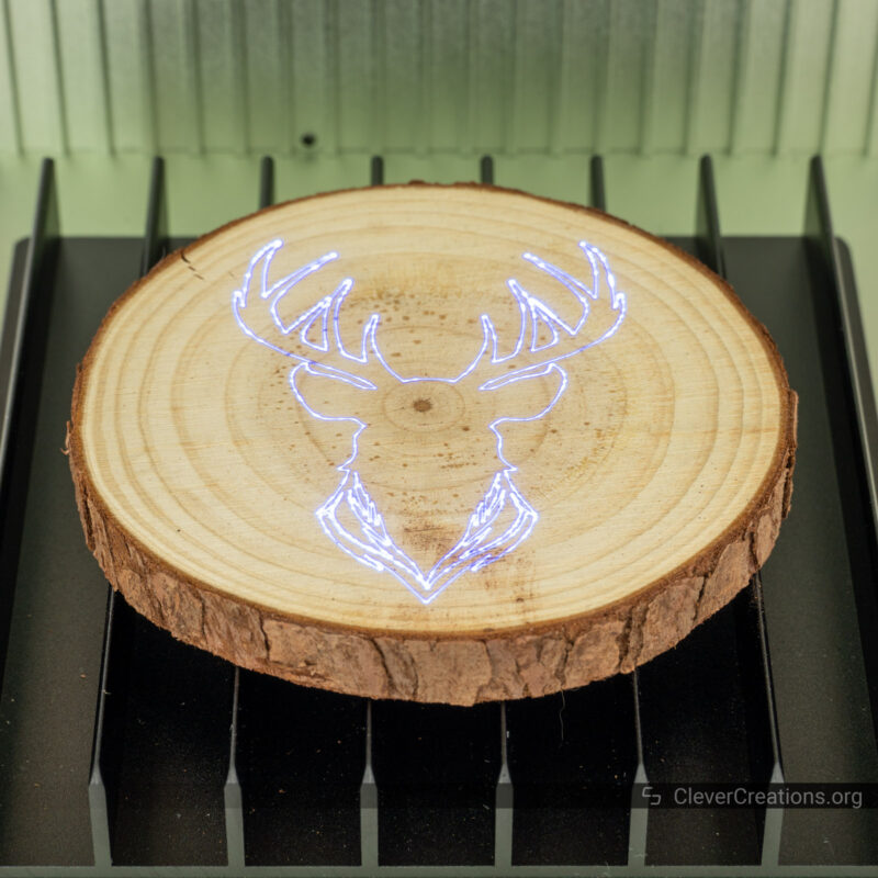 A round wooden coaster with the outline of a deer head in laser light.