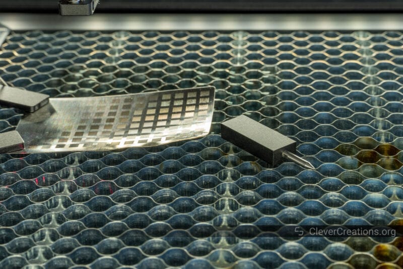 The magnetic touch probe of the xTool S1 placed on a honeycomb panel next to a curved piece of steel.