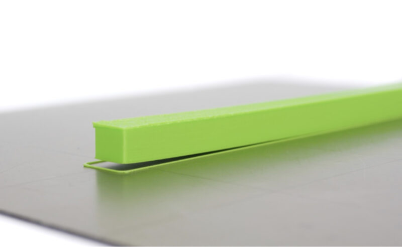 A thin green ABS 3D print with one of its sides lifting off the print bed.
