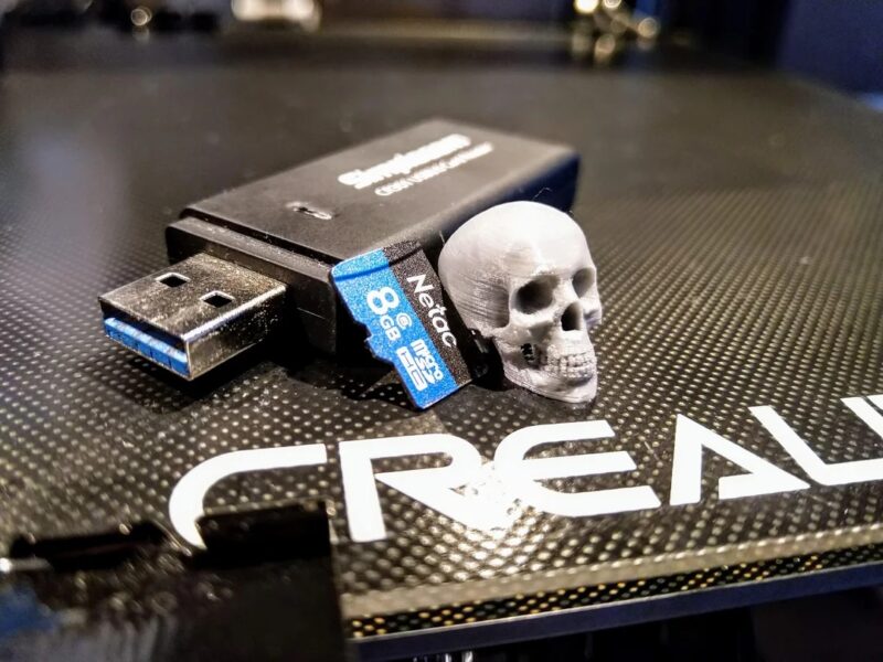 An 8GB Netac Micro-SD card with USB card reader placed on a Creality Ender 3 print surface.