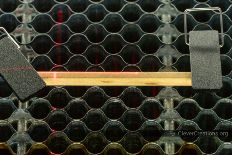 A red laser cross on a pencil that is held in place by clamps on a laser bed.
